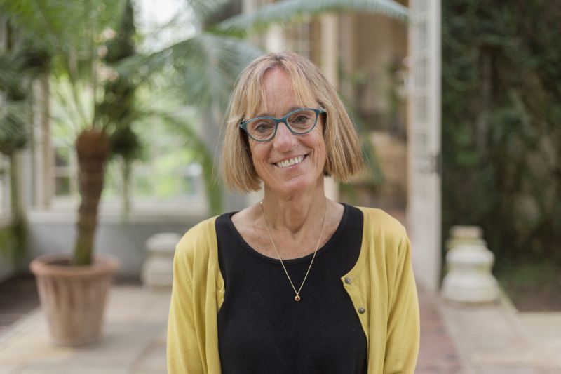 Color photograph of the head and torso of a light skinned woman with blond hair; she stands in an outdoor location, wears glasses, and smiles directly at the camera