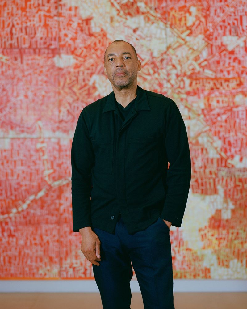 Color photo of a dark skinned man wearing a black blazer and blue pants and standing in front of a red painting
