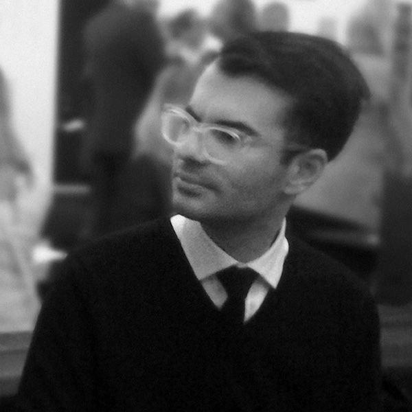 Black and white portrait of Robert Gerard Pietrusko wearing a sweater, collared shirt, and tie, with his torso facing forward but his head in profile