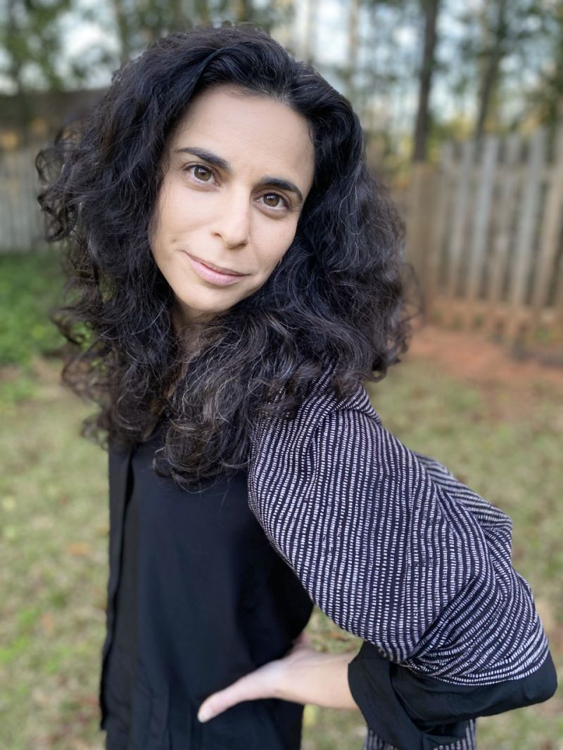 Color photograph of a light skinned woman with dark wavy hair standing in a fenced-in backyard; she stands facing left with her hands on her hips, looking directly into the camera