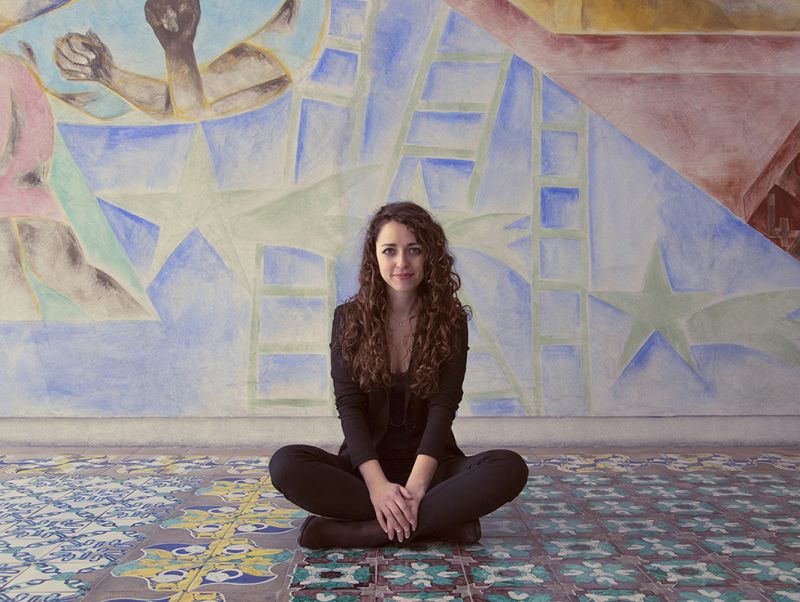Color photograph of Rosa Sessa sitting criss-cross on a colorful tiled floor in front of a colorful wall mural