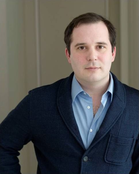Color photo of the head and torso of a light skinned man with dark hair and wearing a dark blue sweater over a light blue shirt