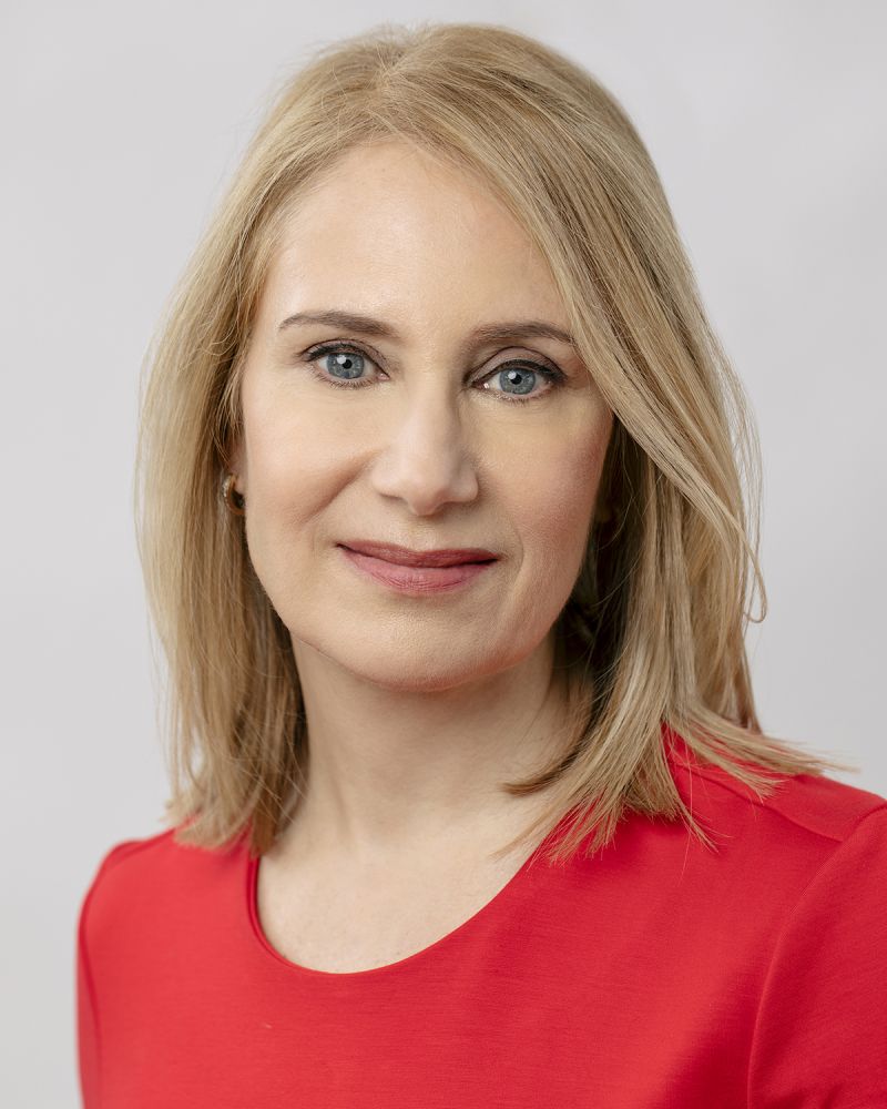 Color photograph of the head and shoulders of Ruth Ben-Ghiat, wearing a red-orange top and looking straight into the camera