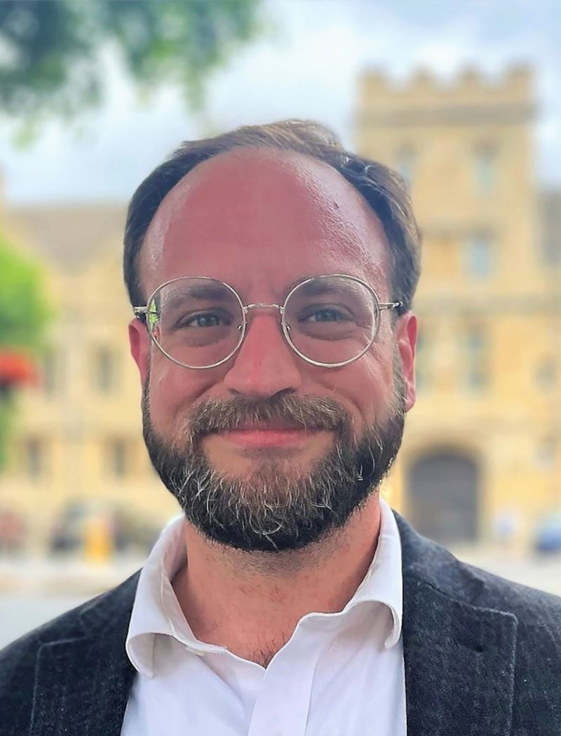 Color photograph of the head and shoulders of a light skinned man with a beard and glasses, smiling at the camera while standing outside with a collegiate building behind him