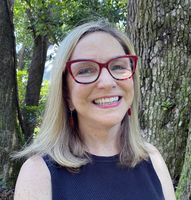 Color photograph of the head and shoulders of a light skinned woman with blond hair wearing large red glasses and smiling at the camera; she stands next to a tree in an outdoor wooden location