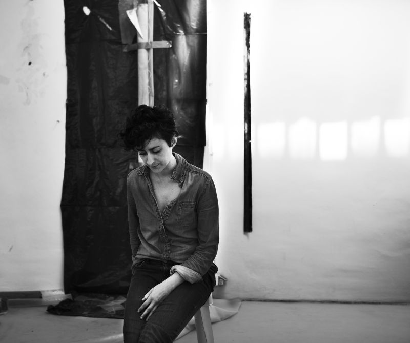 Black and white portrait of the artist Sara Enrico, sitting on a stool in her studio
