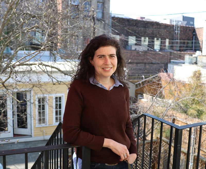 Color photograph of Sarah Nunberg standing on a patio with several New York building backyards behind her