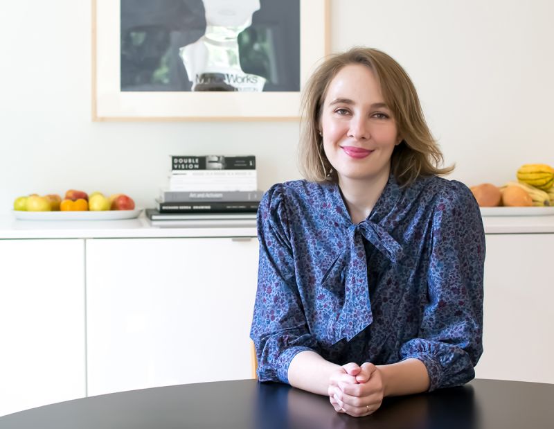 Color photograph of the head and torso of a light skinned woman wearing a patterned blue blouse and smiling at the camera; she sits at a round table in front of a self containing bowls of fruit and a stack of books