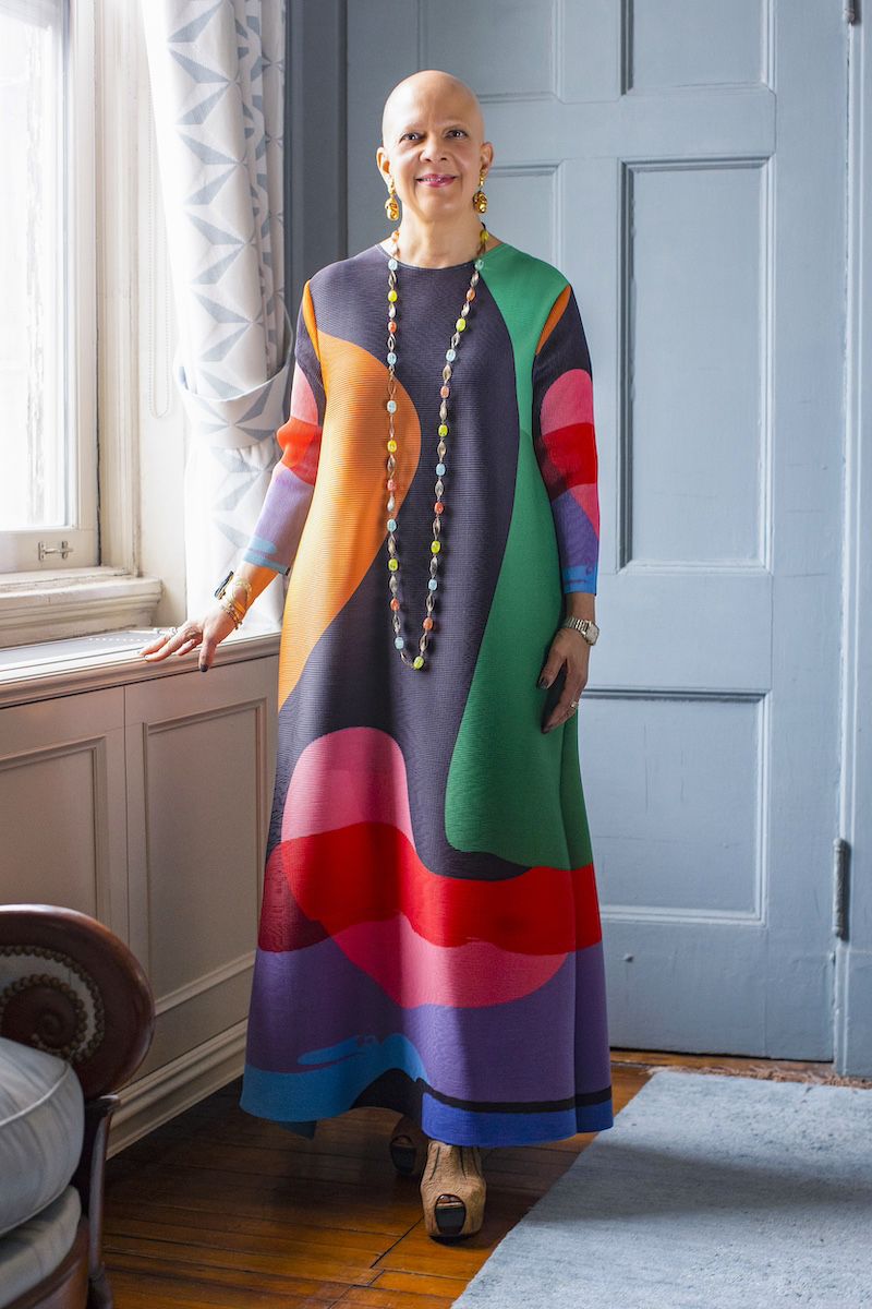 Color photo of a brown skinned woman with a shaved head wearing a flowing colorful dress