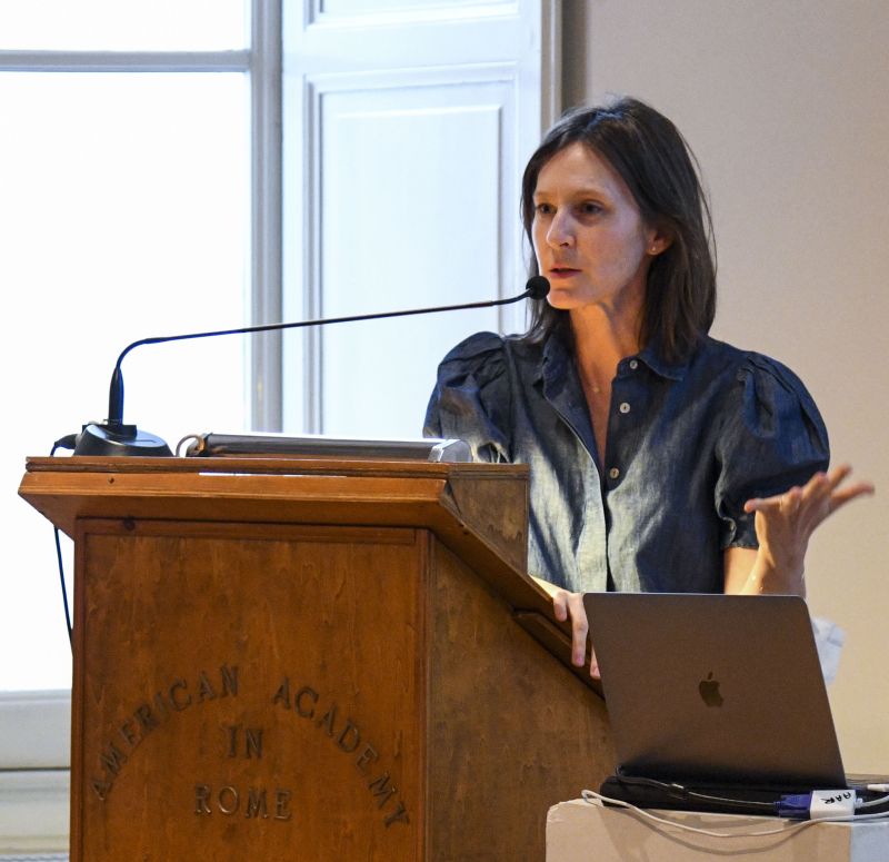 Color photograph of a light skinned brown haired woman standing and speaking at a podium in a lecture room