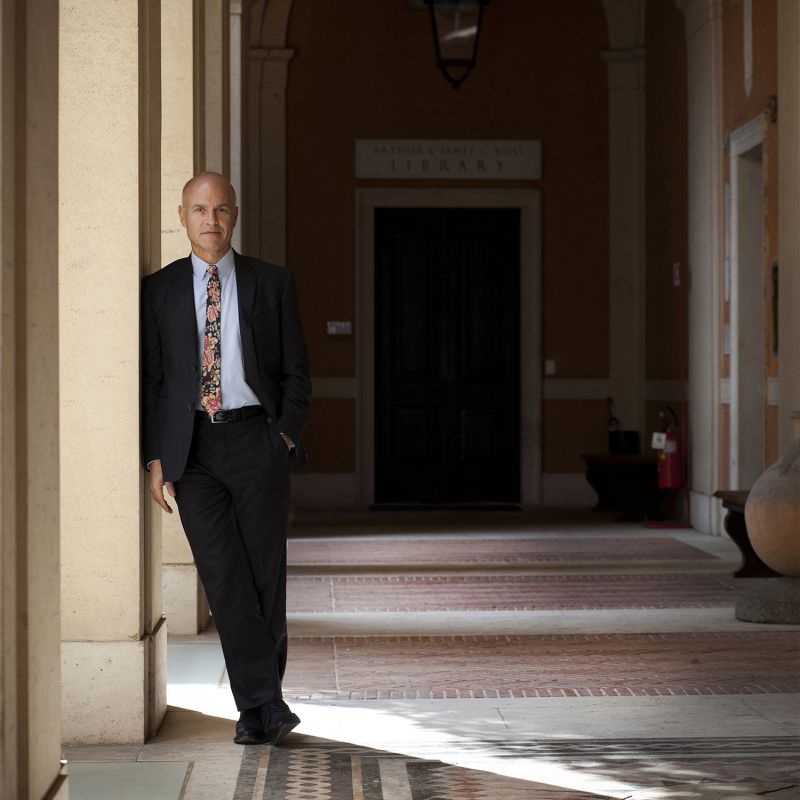 Color photograph of a light skinned man wearing a dark suit and leaning against a column in a Roman cortile