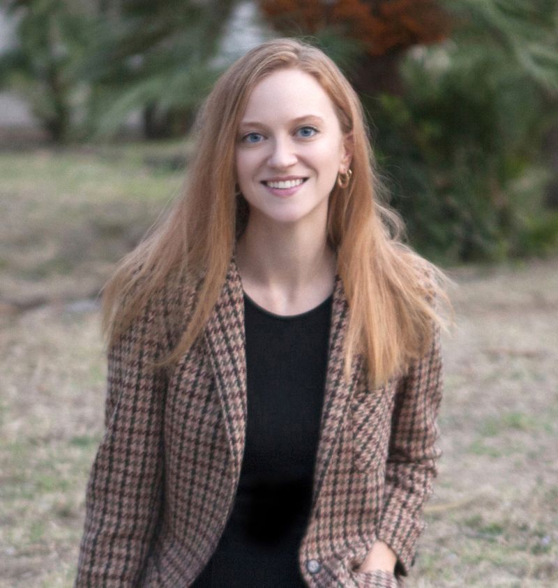 Color photograph of the head and torso of a light skinned woman with red hair smiling at the camera; she is situated in an outdoor natural location and wears a tweed blazer over a black top