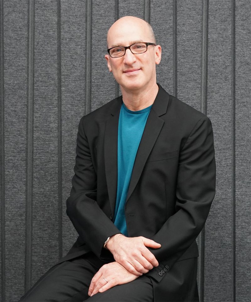 Color photograph of a light skinned man seen from the waist up looking at the camera; he wears glasses and an aqua shirt under a dark blazer