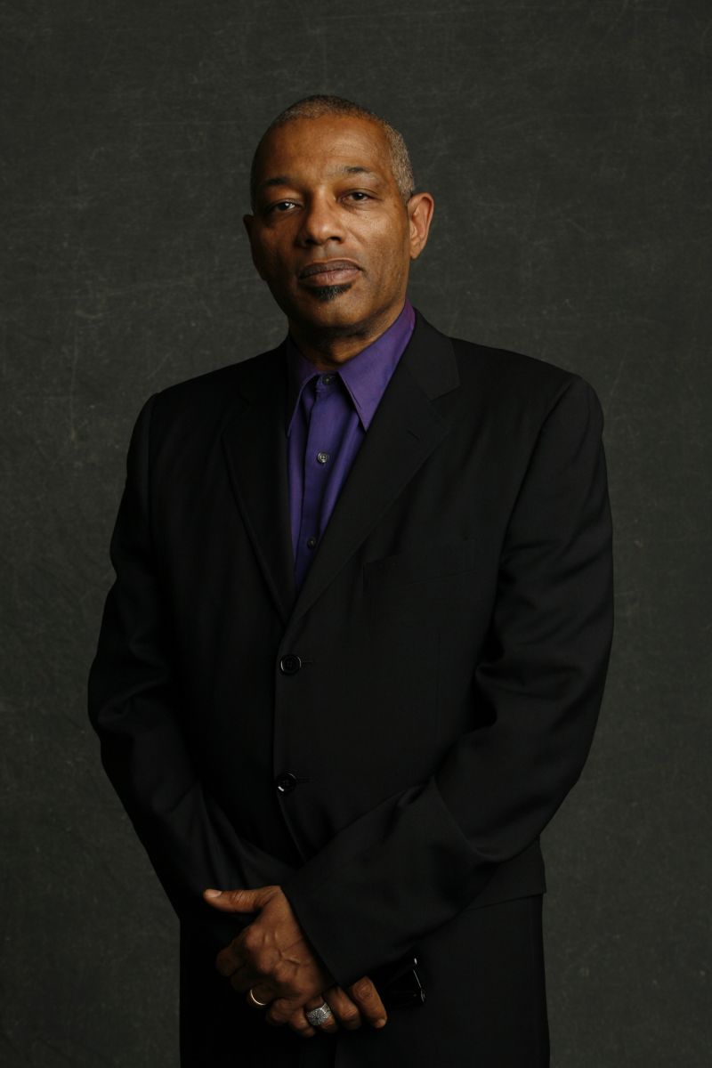 Color photograph of a dark skinned man seen from the waist up; he stands in a photographer's studio and looks directly at the camera