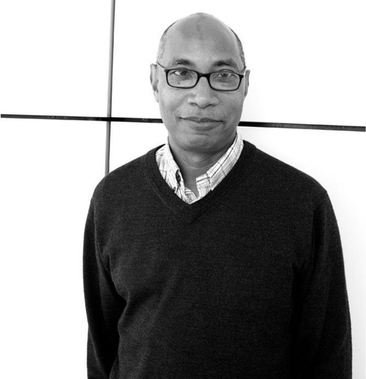 Black and white photograph of the head and torso of a dark skinned man wearing glasses and a sweater over a collared shirt; he looks at the camera with a slight smile