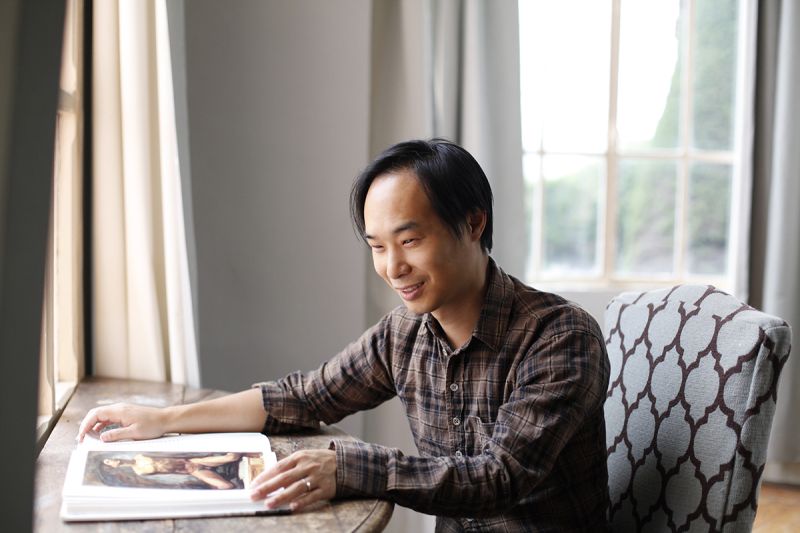 Color photograph of the head and torso of an Asian man wearing a plaid shirt and seated at a desk; he places his hands on the pages of an open book in front of him; light comes into the room from windows in front of and behind him