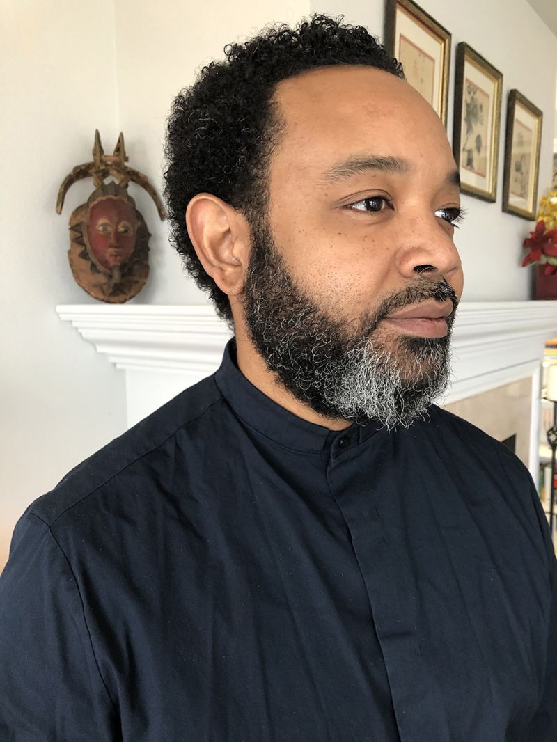 Color photograph of William Villalongo in 3/4 profile, indoors with an African mask on the mantle behind him