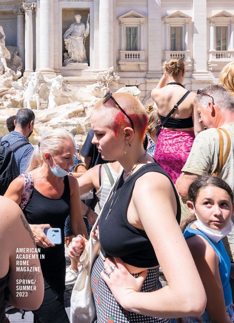 Color photograph of the profile of a young light skinned woman in a black tank top and close cropped hair, seen from the waist up; she stands in front of numerous other tourists in front of the Trevi Fountain in Rome, part of which can be seen in the distance