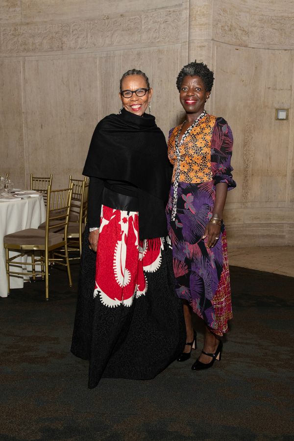 Color photograph of two dark skinned woman at a fundraising gala; on the left the woman wears a black top and black, red, and white skirt; on the right the woman wears an orange, purple, red, and pink patterned dress; both are smiling and looking at the camera