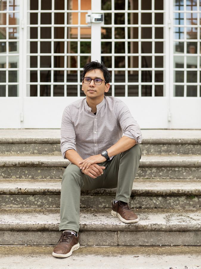 Color photo of an olive skinned man sitting on stone steps in front of a gridded white gate