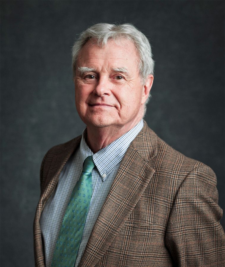 Color photo of an older light skinned man looking at the camera while standing in a portrait studio backdrop