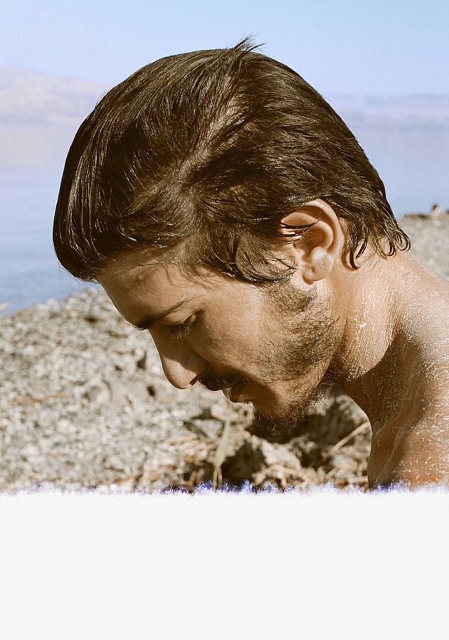 Color photograph of the head of a light skinned man with brown hair seen in profile; behind the head is the shore of the Dead Sea, with rocks and water