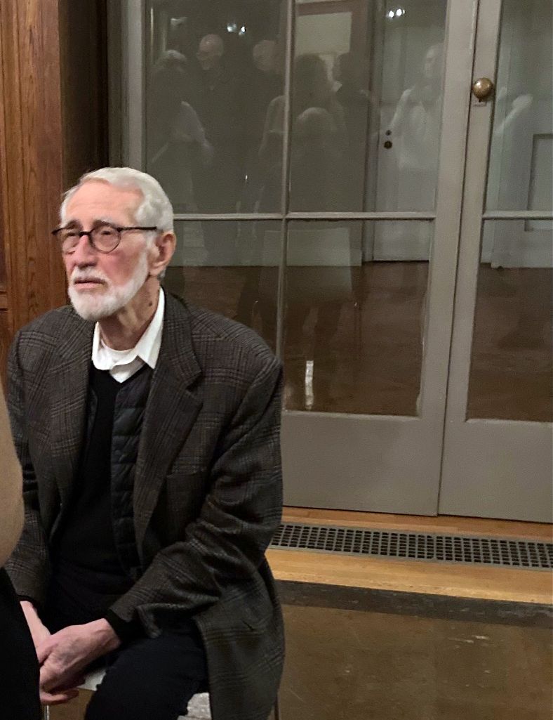 Color photograph of a light skinned man wearing glasses and a white beard sitting in a chair