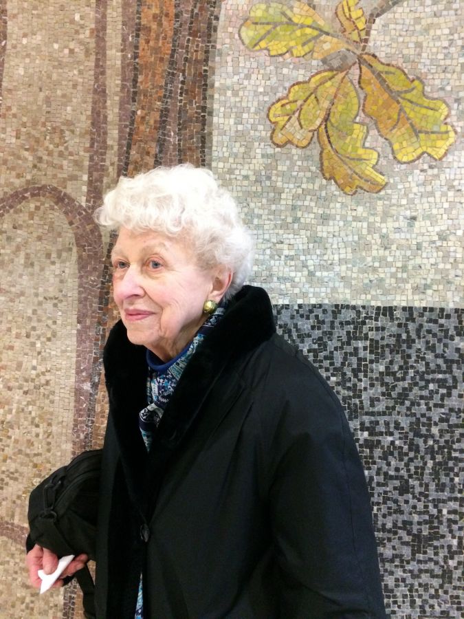 Color photograph of an elderly light skinned woman with white hair in front of a colorful abstract mosaic