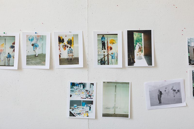 Color photograph of a studio wall that has various color photos of the artist Cy Twombly painting pinned to it in two horizontal rows